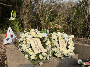 White floral tribute on a stone grave with wooden Japanese inscriptions and a small South Korean flag nearby.