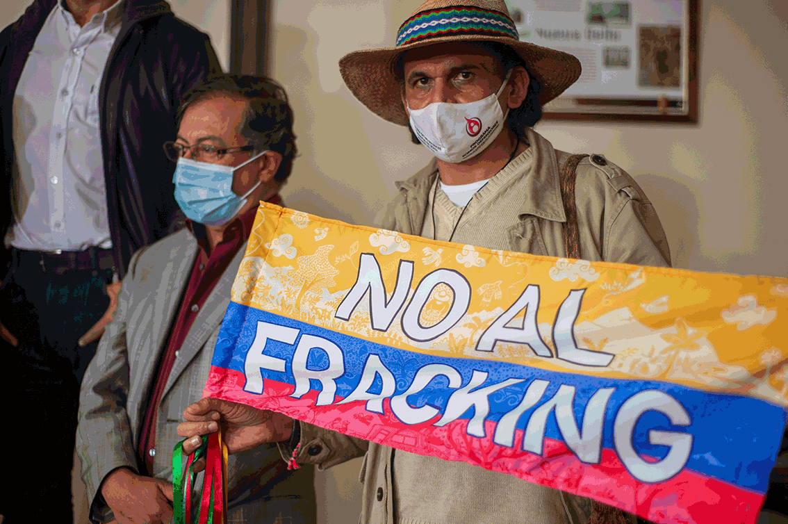 Two men wearing face masks hold a colorful protest banner that reads 'NO ALL FRACKING'.