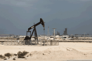 Oil pumpjack in a sandy desert with an industrial facility and power lines in the background, fenced off.