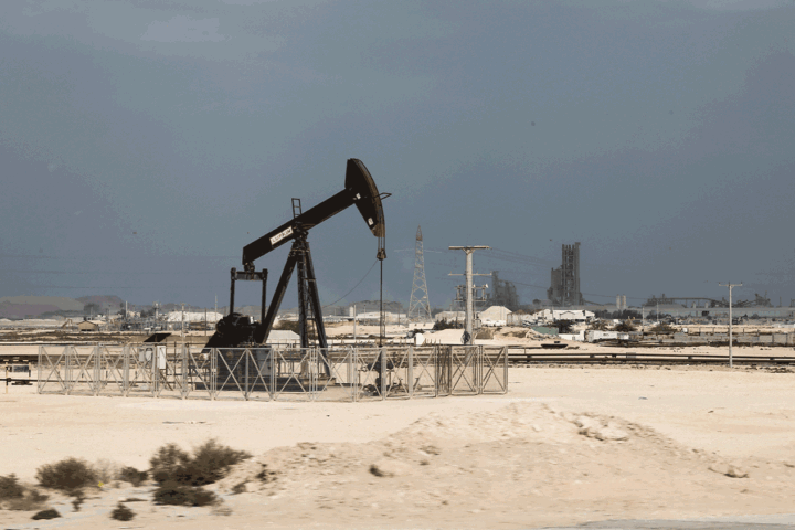 Oil pumpjack in a sandy desert with an industrial facility and power lines in the background, fenced off.