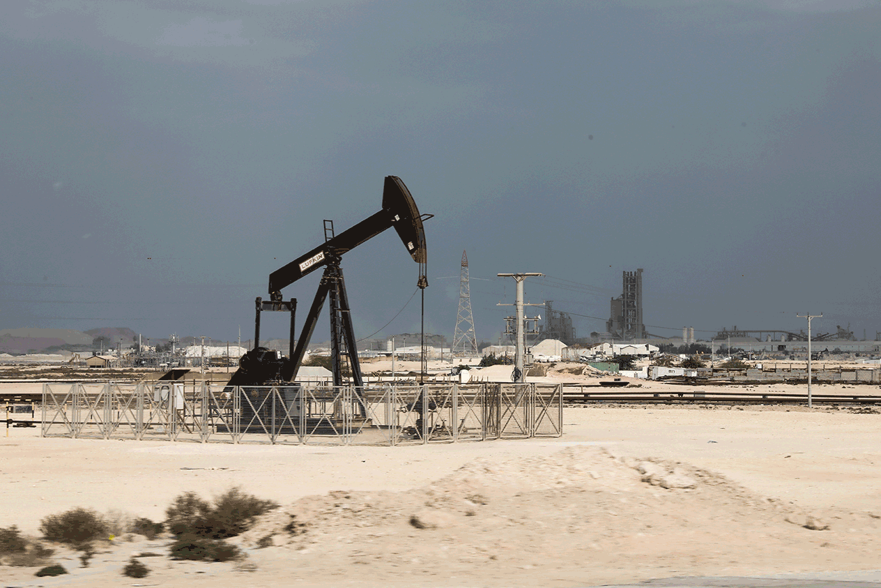 Oil pumpjack in a sandy desert with an industrial facility and power lines in the background, fenced off.