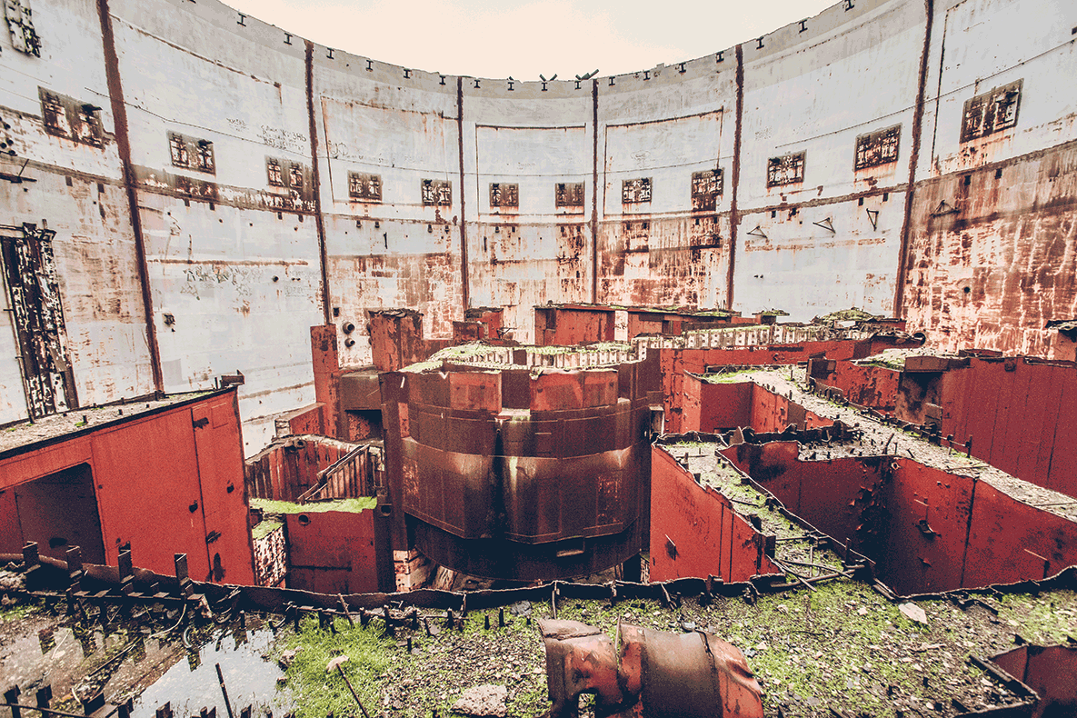Interior of an abandoned industrial site: circular concrete walls, rusted red machinery, and mossy ground visible throughout the scene.