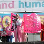 Protest on a stage with people holding bright pink banners and Japanese text behind a speaker at a mic.