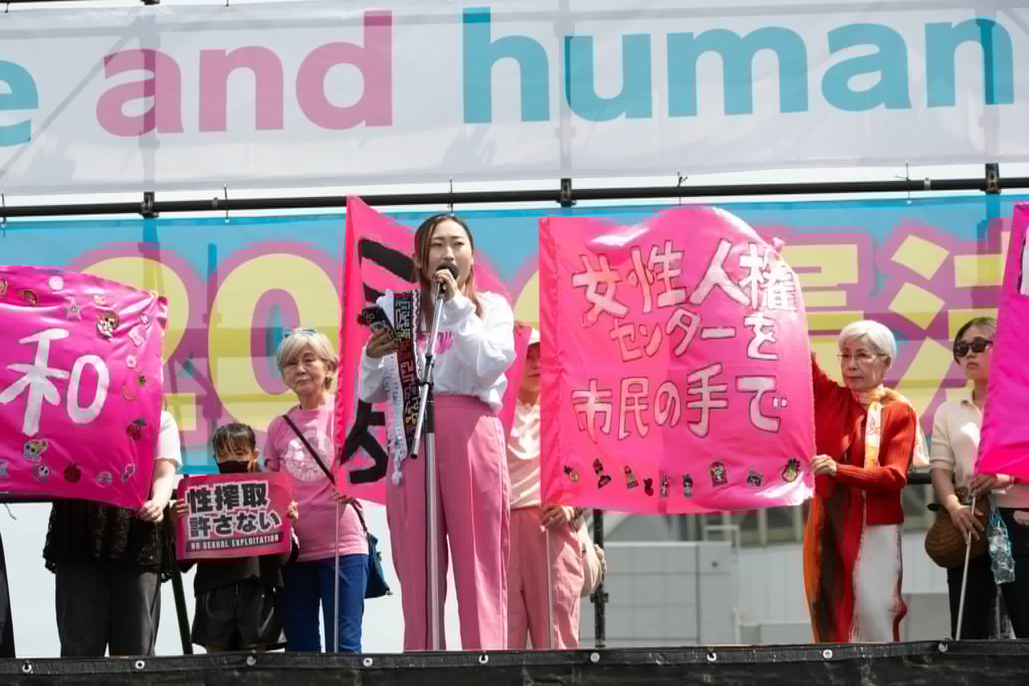 Protest on a stage with people holding bright pink banners and Japanese text behind a speaker at a mic.