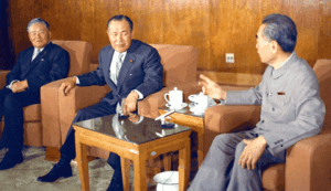 Three men in suits sit in a formal meeting room, coffee cups on a side table between them.