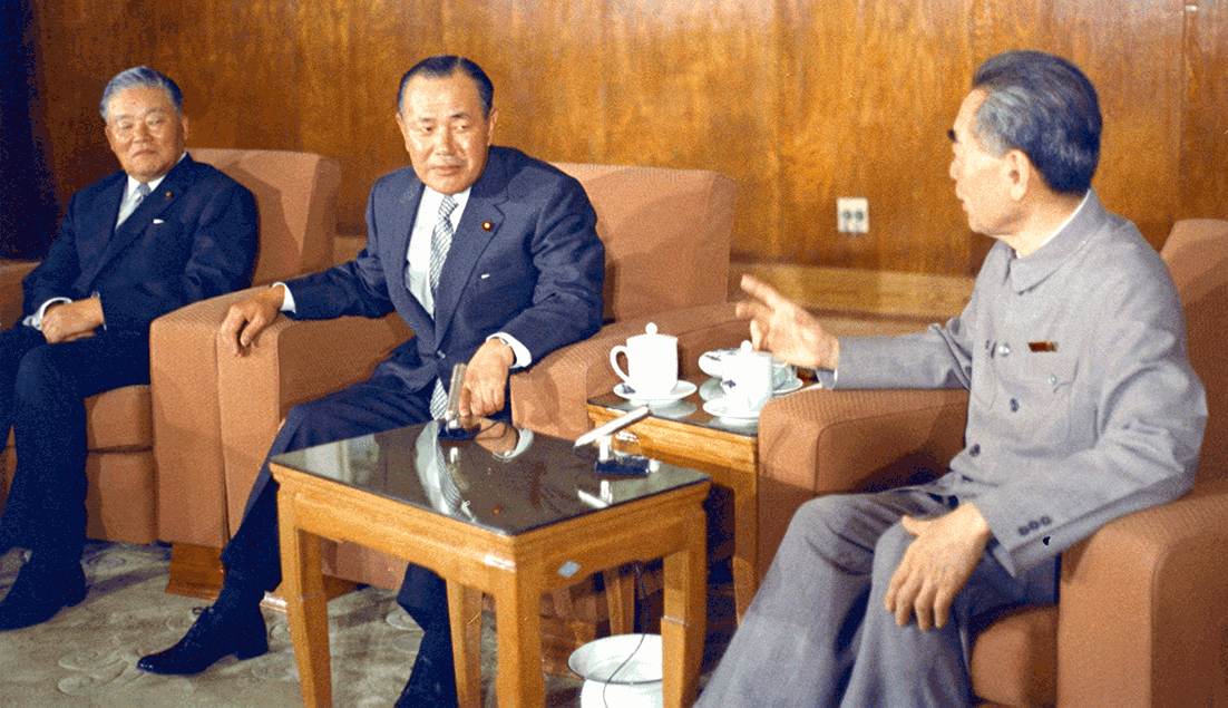 Three men in suits sit in a formal meeting room, coffee cups on a side table between them.