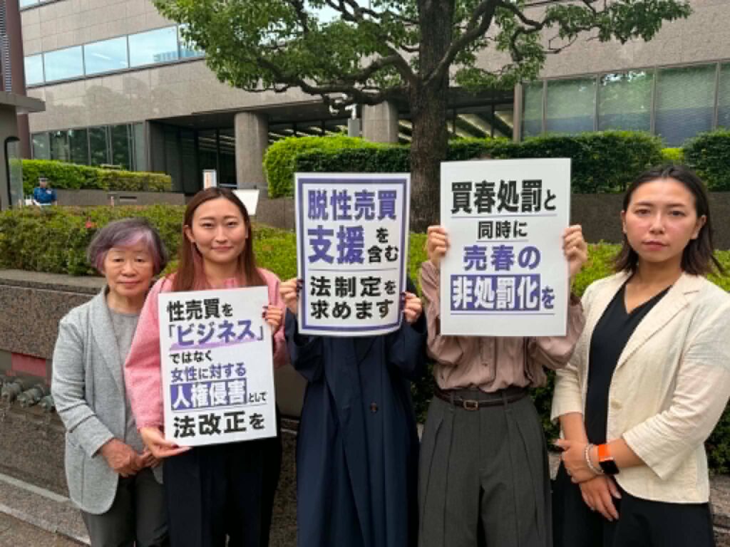 Group of protesters, mostly women, standing in front of a modern building and holding signs with Japanese text.
