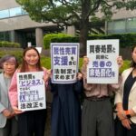 Group of protesters, mostly women, standing in front of a modern building and holding signs with Japanese text.
