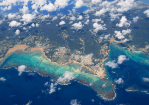 Aerial view of a tropical coastline with a small town along white-sand beaches and turquoise coral-reef waters. Clouds drift above the scene.
