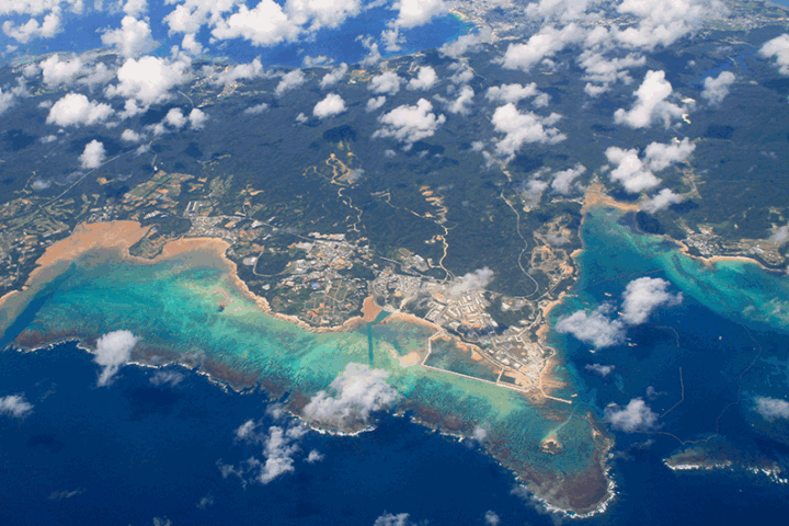 Aerial view of a tropical coastline with a small town along white-sand beaches and turquoise coral-reef waters. Clouds drift above the scene.