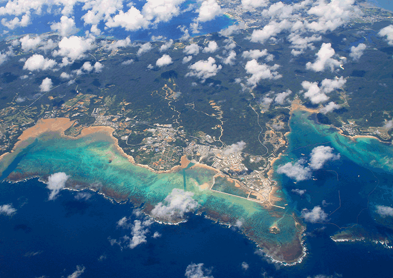 Aerial view of a tropical coastline with a small town along white-sand beaches and turquoise coral-reef waters. Clouds drift above the scene.