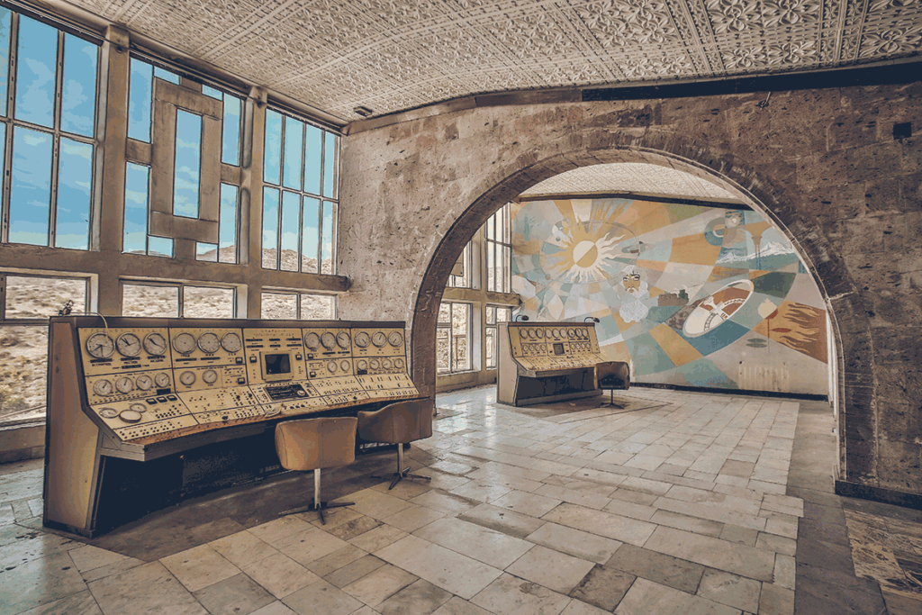 Abandoned control room with vintage panels and chairs beneath a decorative ceiling and a mural through an arched opening.