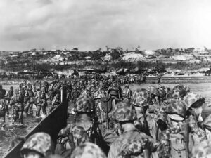 Soldiers in camouflage and helmets disembark from a landing craft onto a sandy beach during a military operation.