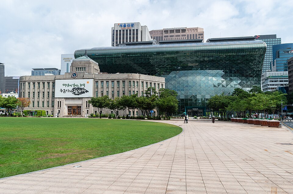 Public square with a large modern glass building behind a historic stone building, a green lawn, and a wide tiled promenade with trees and a few pedestrians.