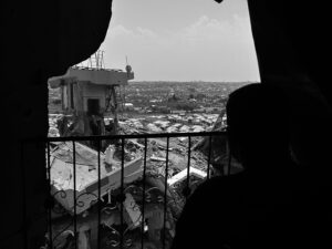Silhouetted figures stand behind a barred balcony, overlooking a destroyed cityscape with rubble and ruined buildings under a bright sky.