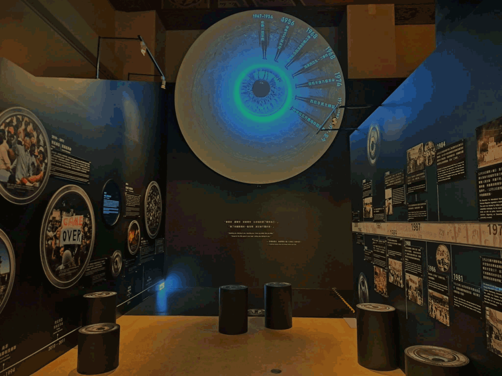 Large circular wall display resembling an eye with a blue-green center, timeline labels around the edge in a dark museum gallery with text panels nearby.
