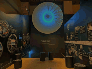 Large circular wall display resembling an eye with a blue-green center, timeline labels around the edge in a dark museum gallery with text panels nearby.