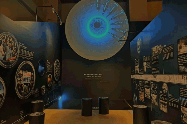Large circular wall display resembling an eye with a blue-green center, timeline labels around the edge in a dark museum gallery with text panels nearby.