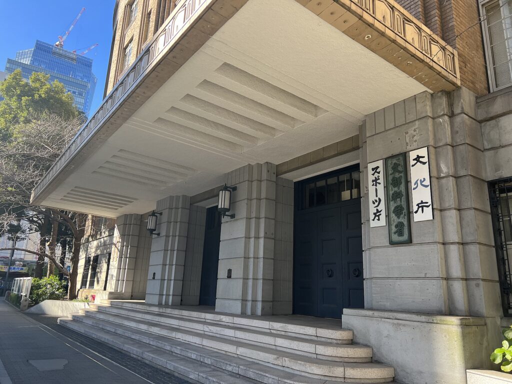 Entrance to a stone building with a large white overhang, concrete steps, and vertical Japanese signs beside dark doors; urban street visible to the left.