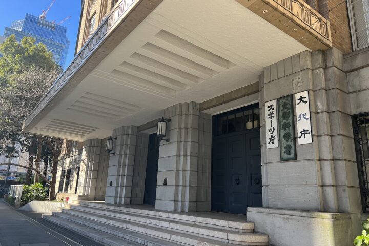 Entrance to a stone building with a large white overhang, concrete steps, and vertical Japanese signs beside dark doors; urban street visible to the left.