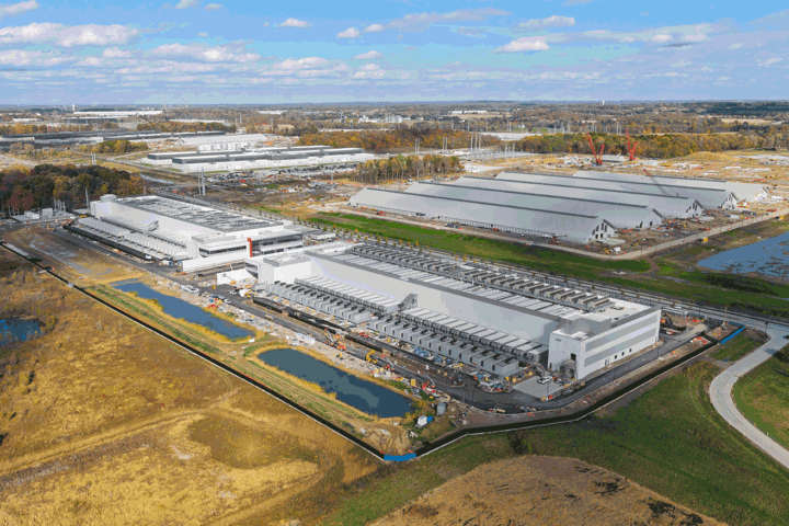 Aerial view of a large industrial complex with multiple white warehouses under construction, cranes, and surrounding fields.