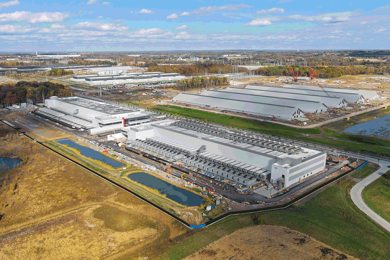 Aerial view of a large industrial complex with multiple white warehouses under construction, cranes, and surrounding fields.