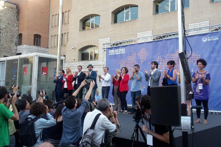 Speakers on a stage clap as photographers in front capture the event near a blue Fearless Cities banner in Barcelona.