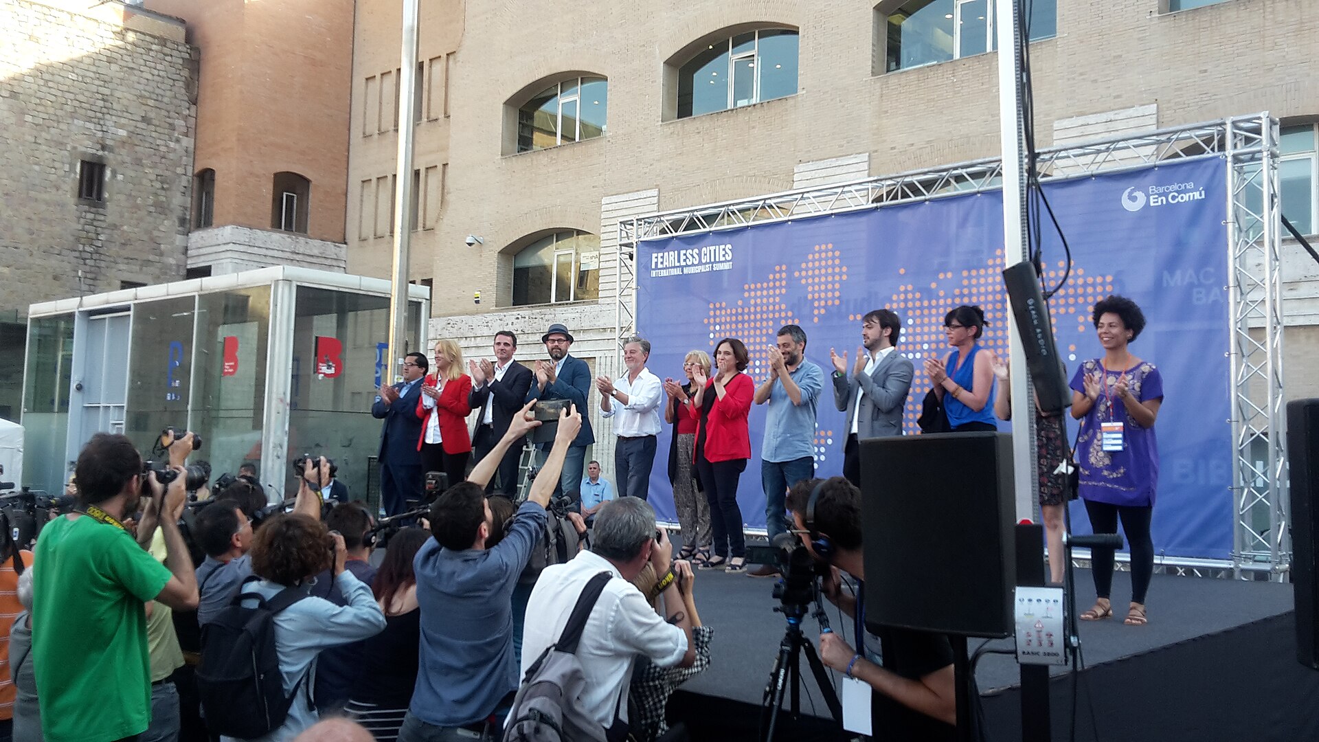 Speakers on a stage clap as photographers in front capture the event near a blue Fearless Cities banner in Barcelona.
