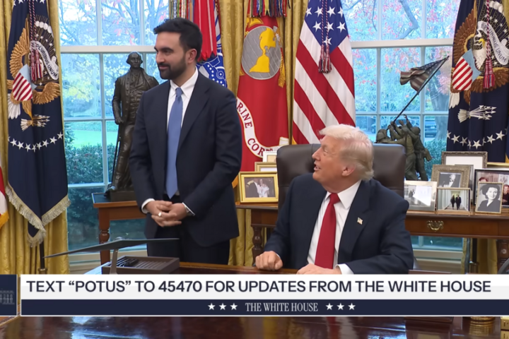In the Oval Office, a man in a dark suit stands beside a seated man in a suit with a red tie; behind them are US flags, a large desk, and framed photos, with a news ticker at the bottom saying “TEXT POTUS TO 45470 FOR UPDATES FROM THE WHITE HOUSE.”