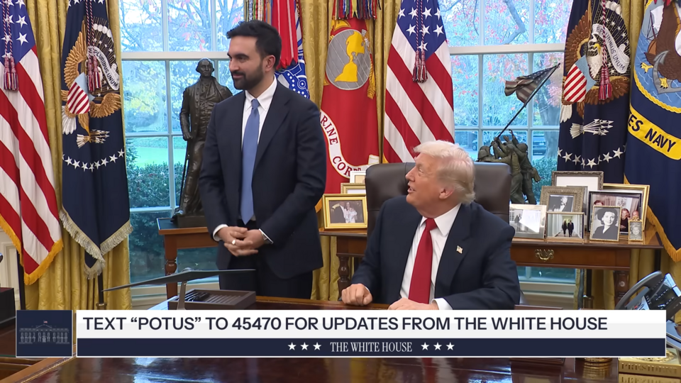 In the Oval Office, a man in a dark suit stands beside a seated man in a suit with a red tie; behind them are US flags, a large desk, and framed photos, with a news ticker at the bottom saying “TEXT POTUS TO 45470 FOR UPDATES FROM THE WHITE HOUSE.”