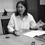 Woman in a white shirt gestures while discussing at a conference table with papers and a patterned pouch.