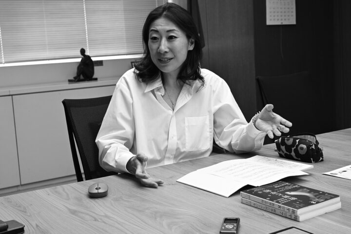 Woman in a white shirt gestures while discussing at a conference table with papers and a patterned pouch.