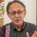 Older man with red glasses speaking, wearing a dark gray shirt, gesturing with his hand; notes pinned on a corkboard in the background.