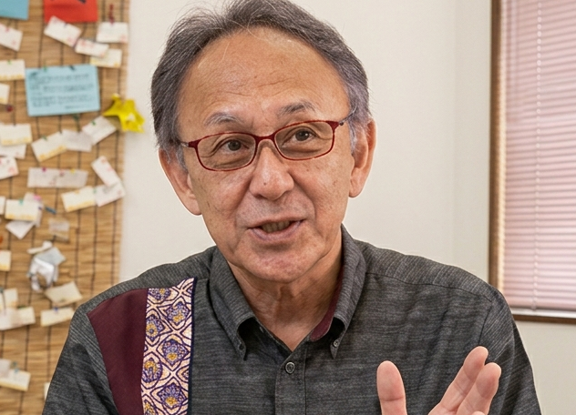 Older man with red glasses speaking, wearing a dark gray shirt, gesturing with his hand; notes pinned on a corkboard in the background.