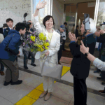 Smiling woman in a white suit waves and holds a bouquet of flowers as photographers and onlookers gather outside a building.