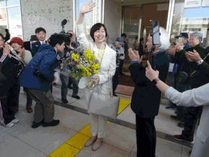 Smiling woman in a white suit waves and holds a bouquet of flowers as photographers and onlookers gather outside a building.