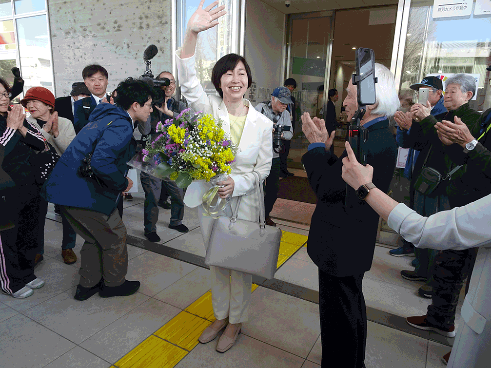 Smiling woman in a white suit waves and holds a bouquet of flowers as photographers and onlookers gather outside a building.
