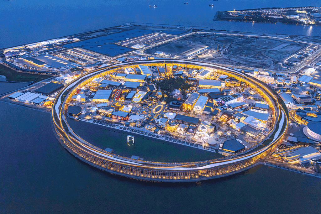 Aerial view of a circular waterfront shopping complex lit up at night on a man-made island.