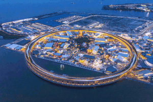 Aerial view of a circular waterfront shopping complex lit up at night on a man-made island.